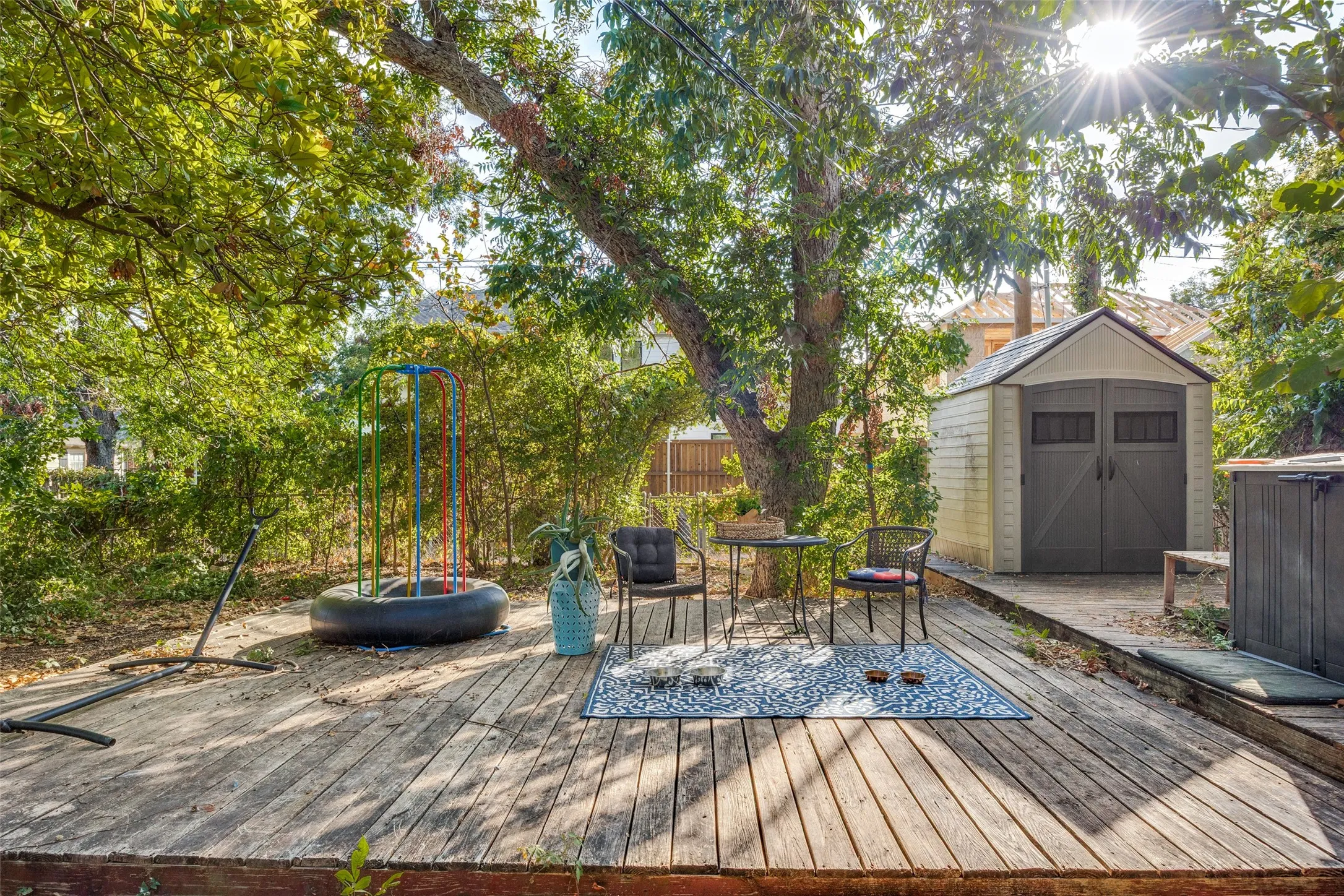 Wood-deck patio area featuring 2 storage sheds with double doors and a pitched roof, mature trees, and dense perimeter foliage