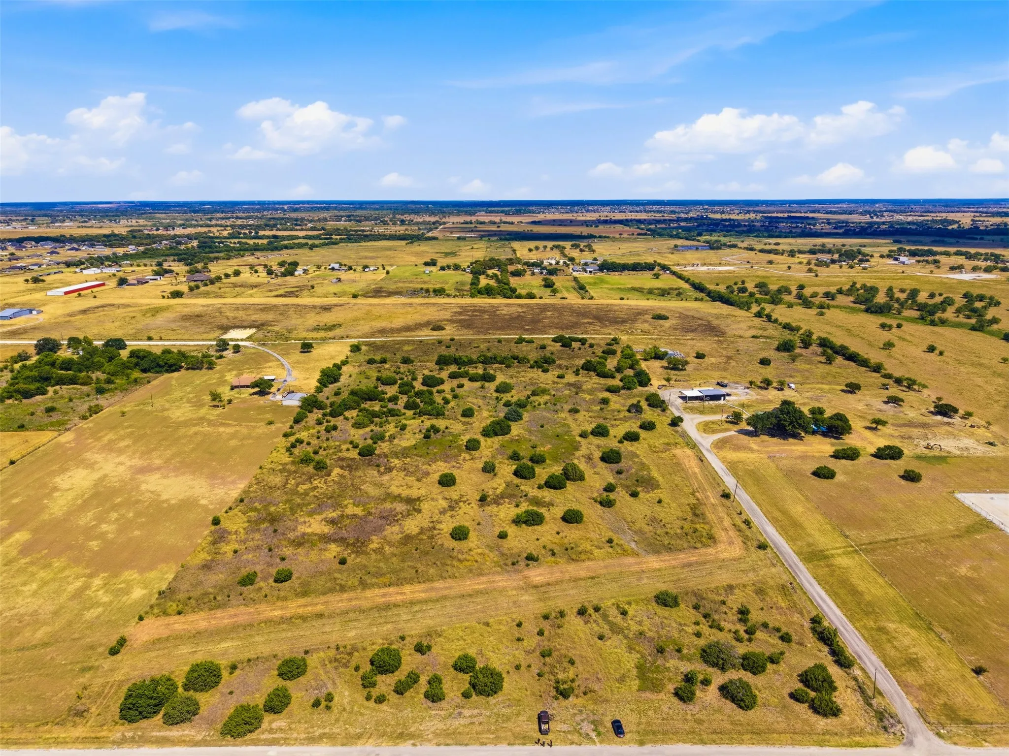 Expansive rural landscape with scattered trees, a paved road, and distant structures under a clear sky