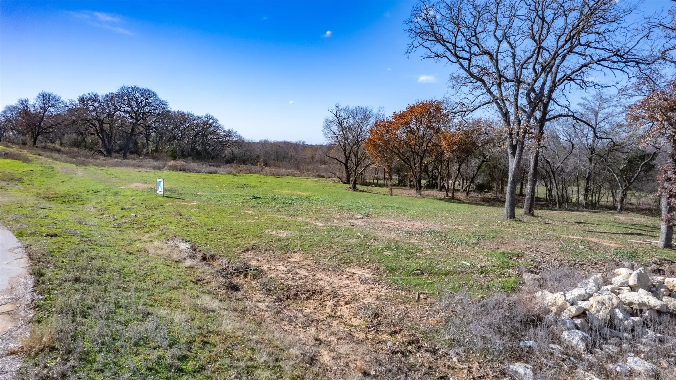 Expansive natural landscape featuring varied terrain, a clear blue sky, mature trees, and a visible stone pile