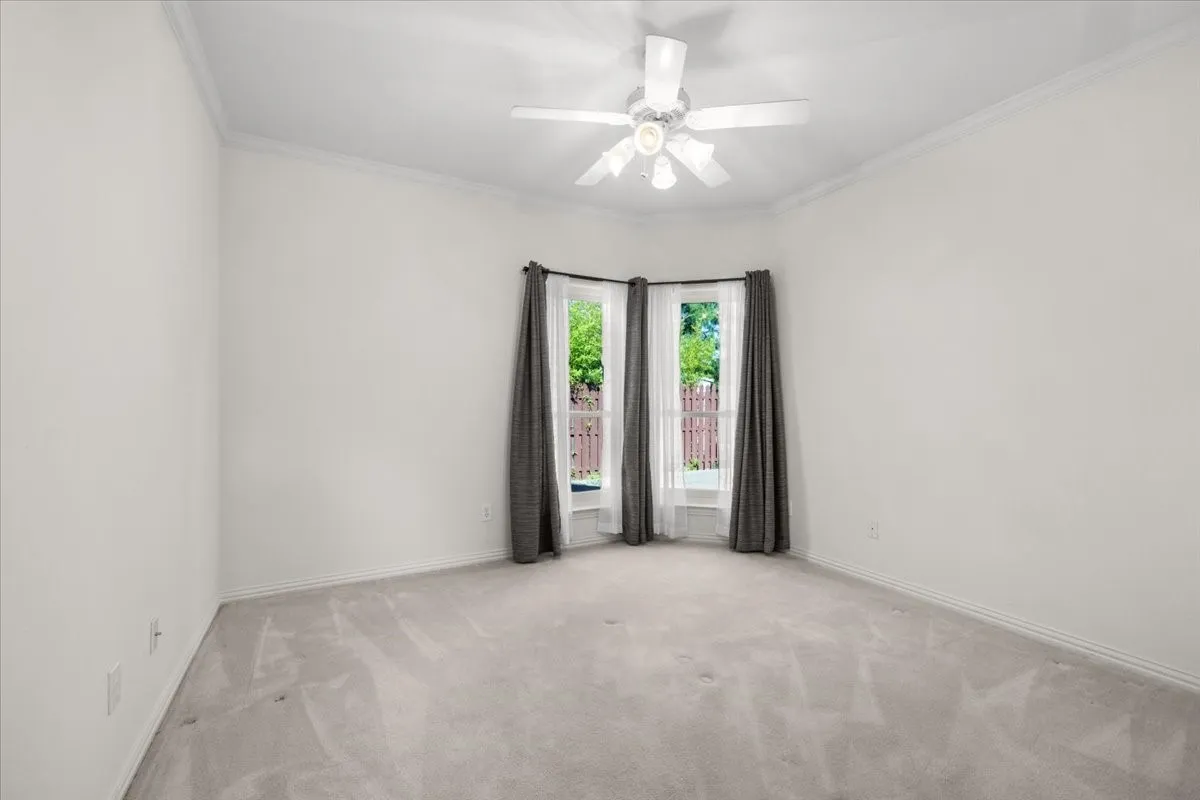 Bay window with outdoor scenery, ceiling fan with light kit, crown molding, and light-colored carpeting