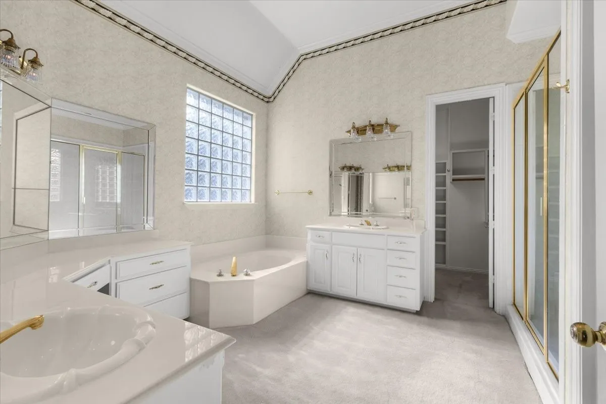 Dual vanity with white cabinetry, glass block window, built-in soaking tub, and shower enclosure with gold-tone framing