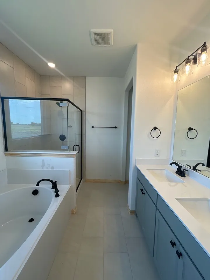 Dual vanity with integrated sinks and black fixtures, a built-in soaking tub, a glass-enclosed shower with tiled walls, and light-toned tile flooring