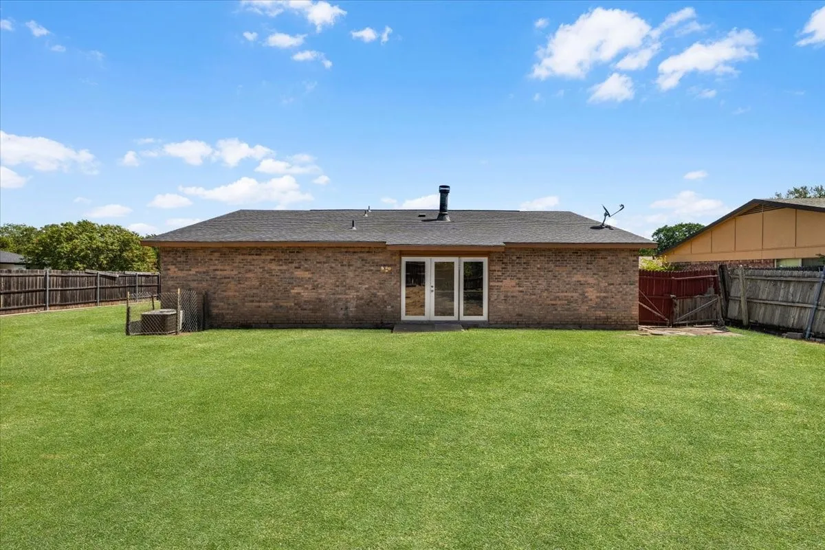 Expansive green lawn, brick exterior, dark shingle roof, double patio doors, and a wood privacy fence