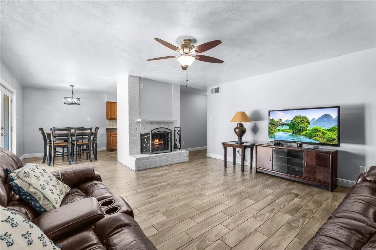 Expansive living area featuring wood-finish flooring, a white brick fireplace, and a ceiling fan