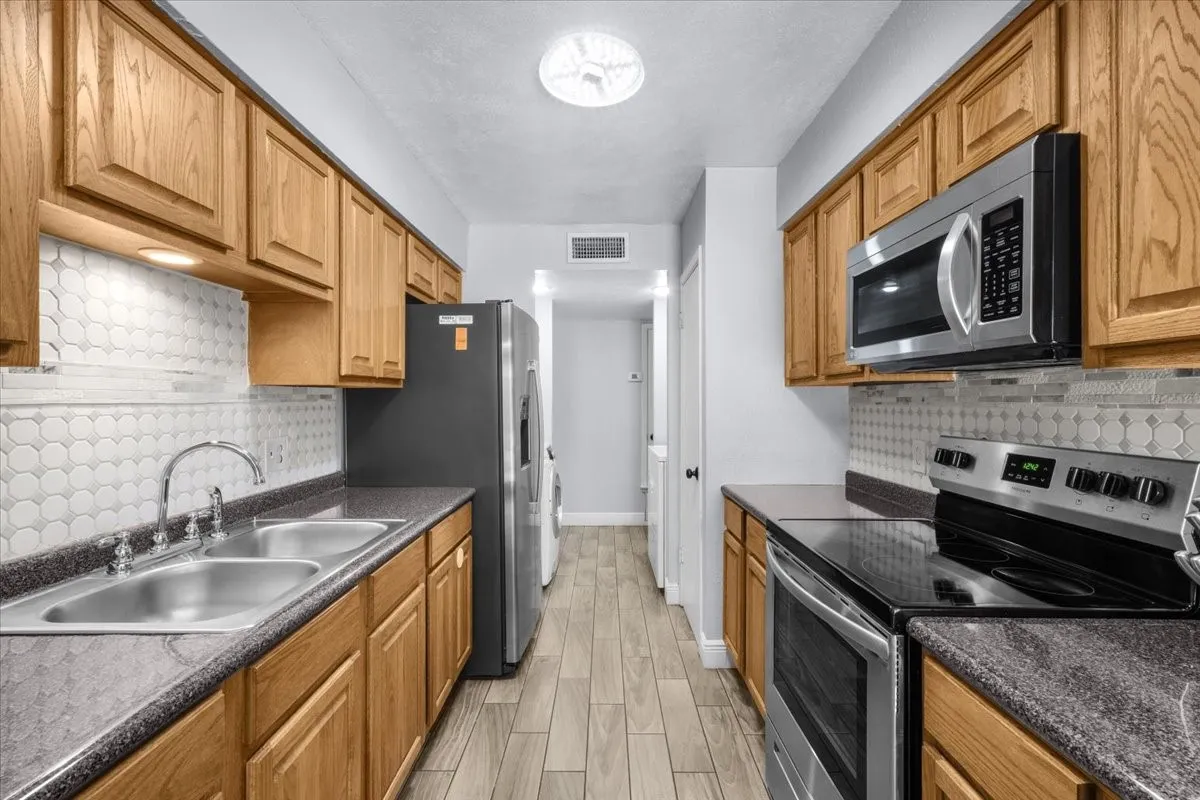 Kitchen featuring wood cabinetry, stainless steel appliances, dual basin sink, patterned tile backsplash, and wood-finish flooring