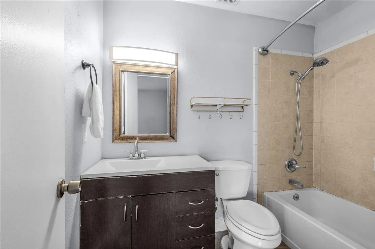 Vanity with integrated sink, rectangular framed mirror, modern wall-mounted light fixture, white bathtub, and tiled shower surround