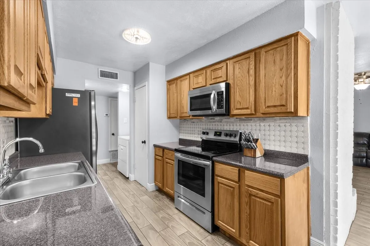 Galley kitchen featuring wood-finish cabinetry, dark countertops, stainless steel appliances, and a tiled backsplash
