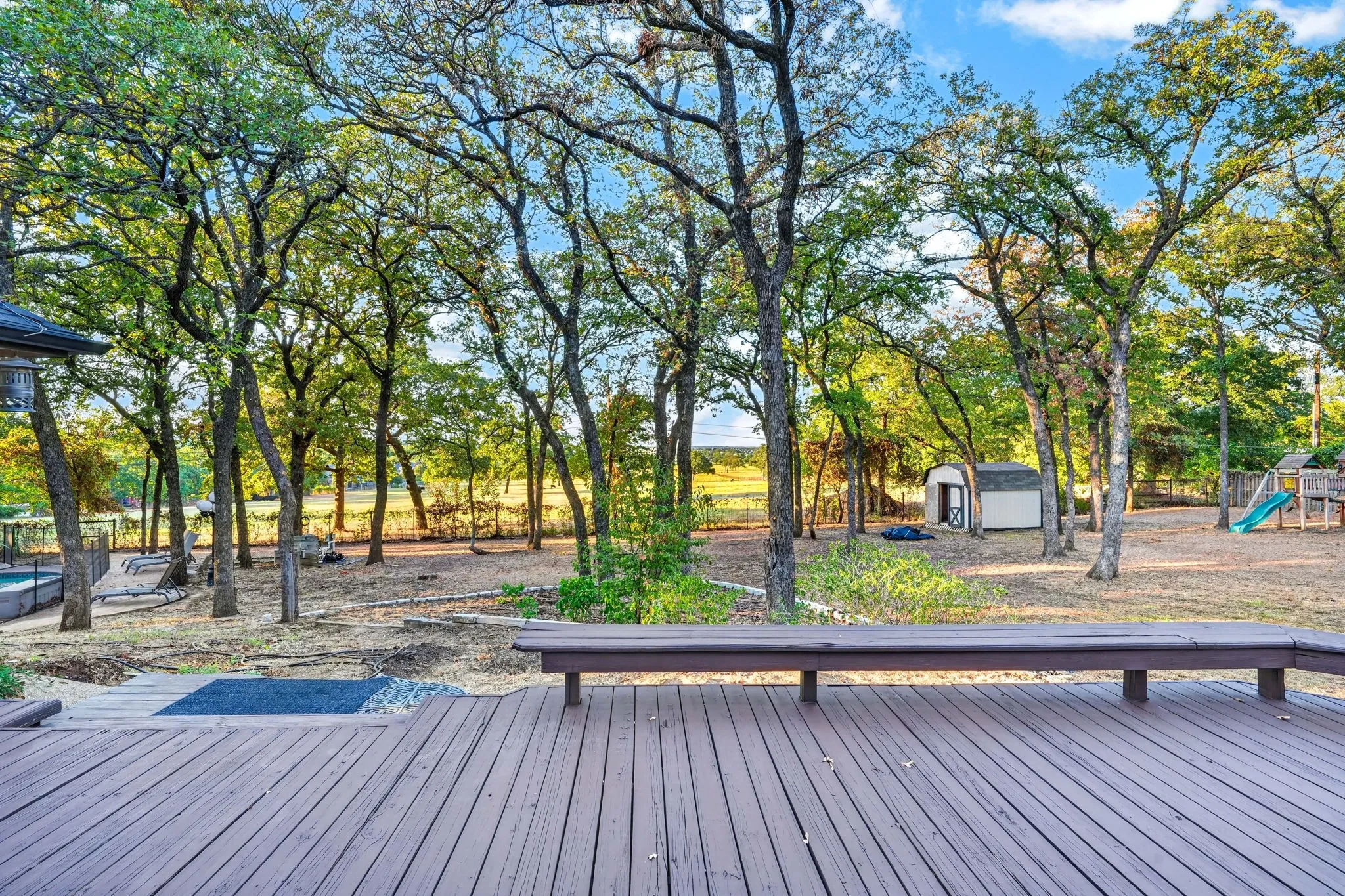 Expansive backyard featuring a composite deck, built-in deck seating, mature trees, a storage shed, and a play structure with view of open land beyond