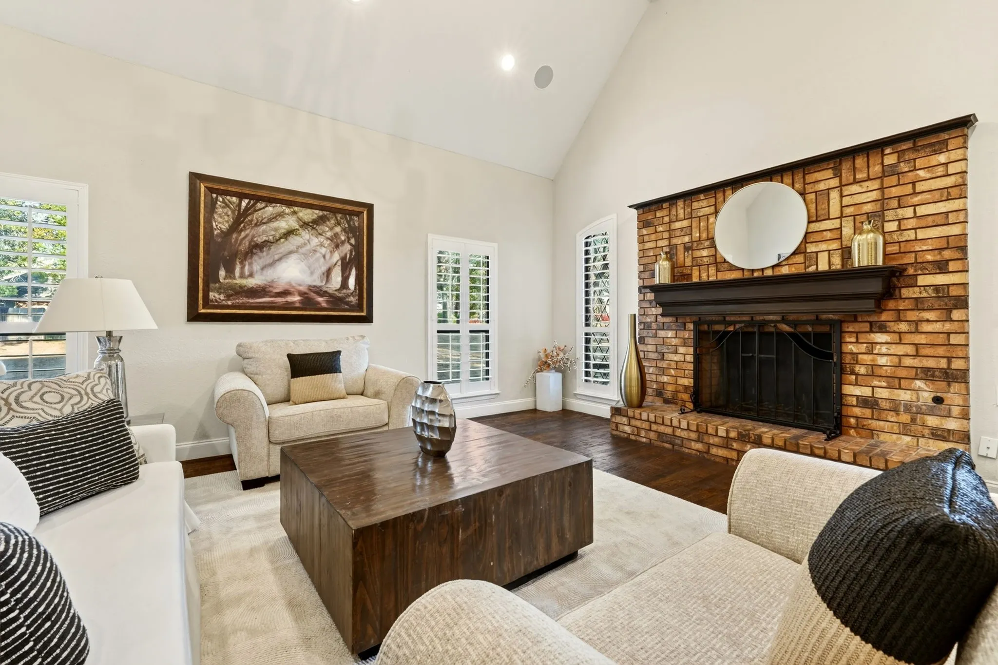 Brick fireplace with dark wood mantel, vaulted ceiling, recessed lighting, dark wood-finish flooring, and white plantation shutters