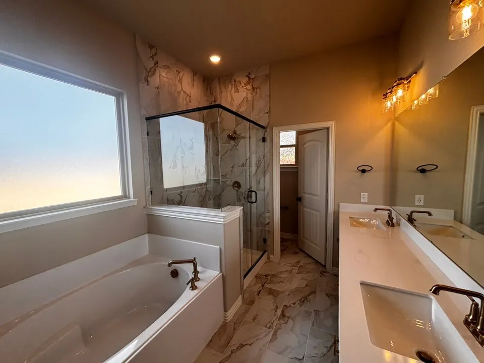 Spacious bathroom featuring a built-in bathtub, glass-enclosed shower with tile surround, double vanity with integrated sinks, and bronze-finish fixtures