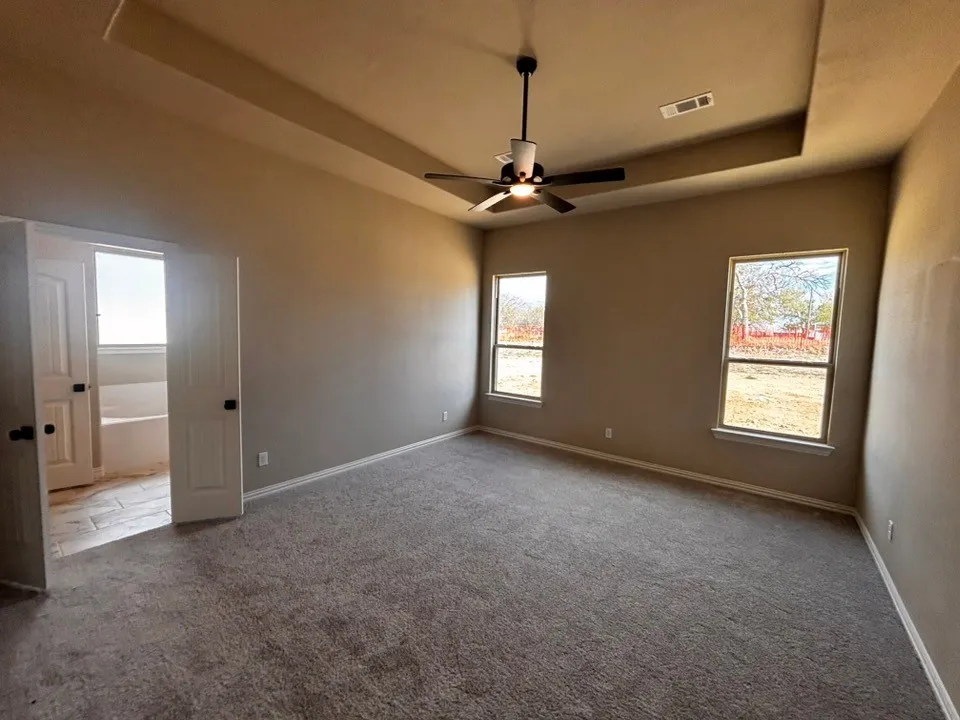 Tray ceiling with a ceiling fan, two windows, carpeted flooring, and a double door entry to an adjoining room with a built-in tub