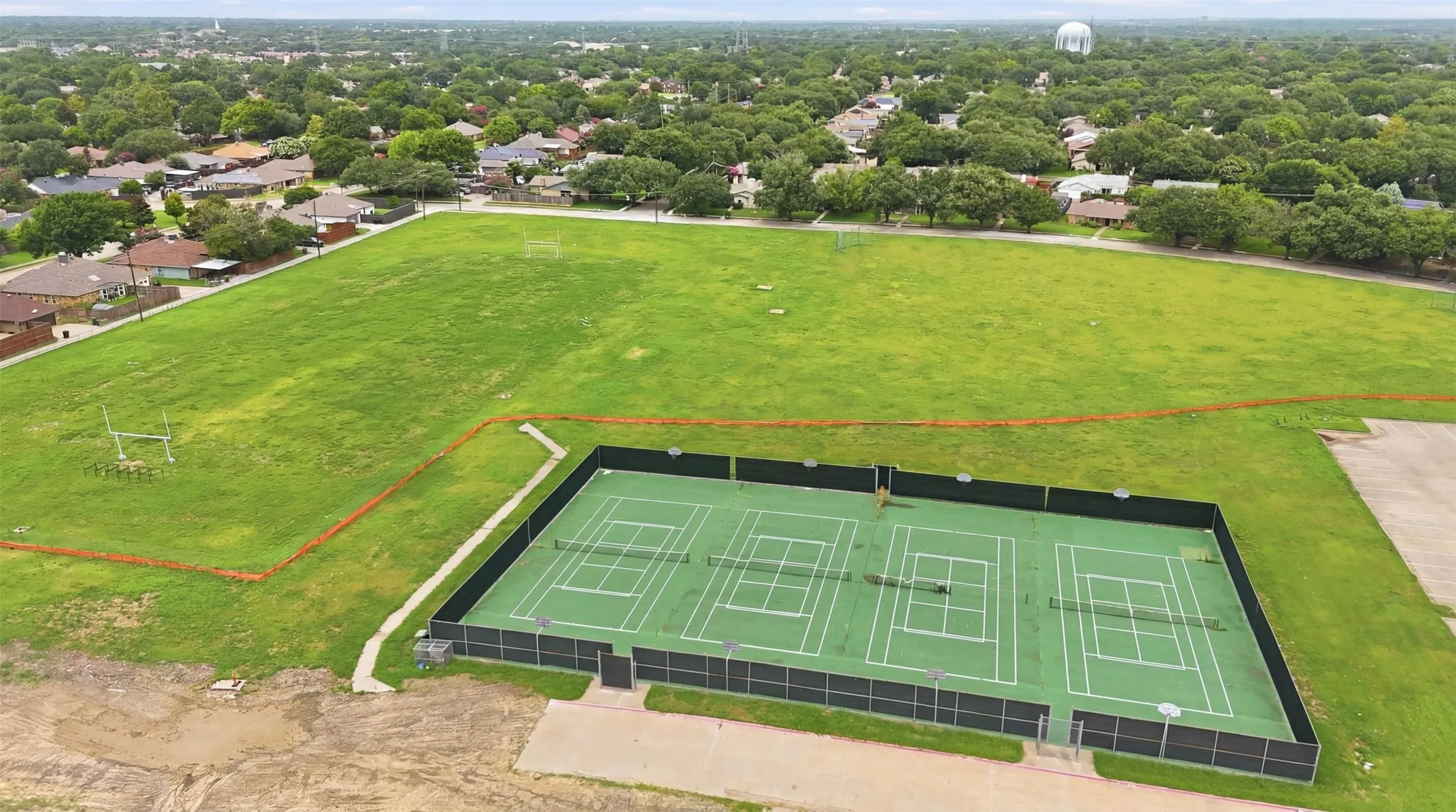 Three fenced tennis courts bordered by a grass field and residential areas with mature trees
