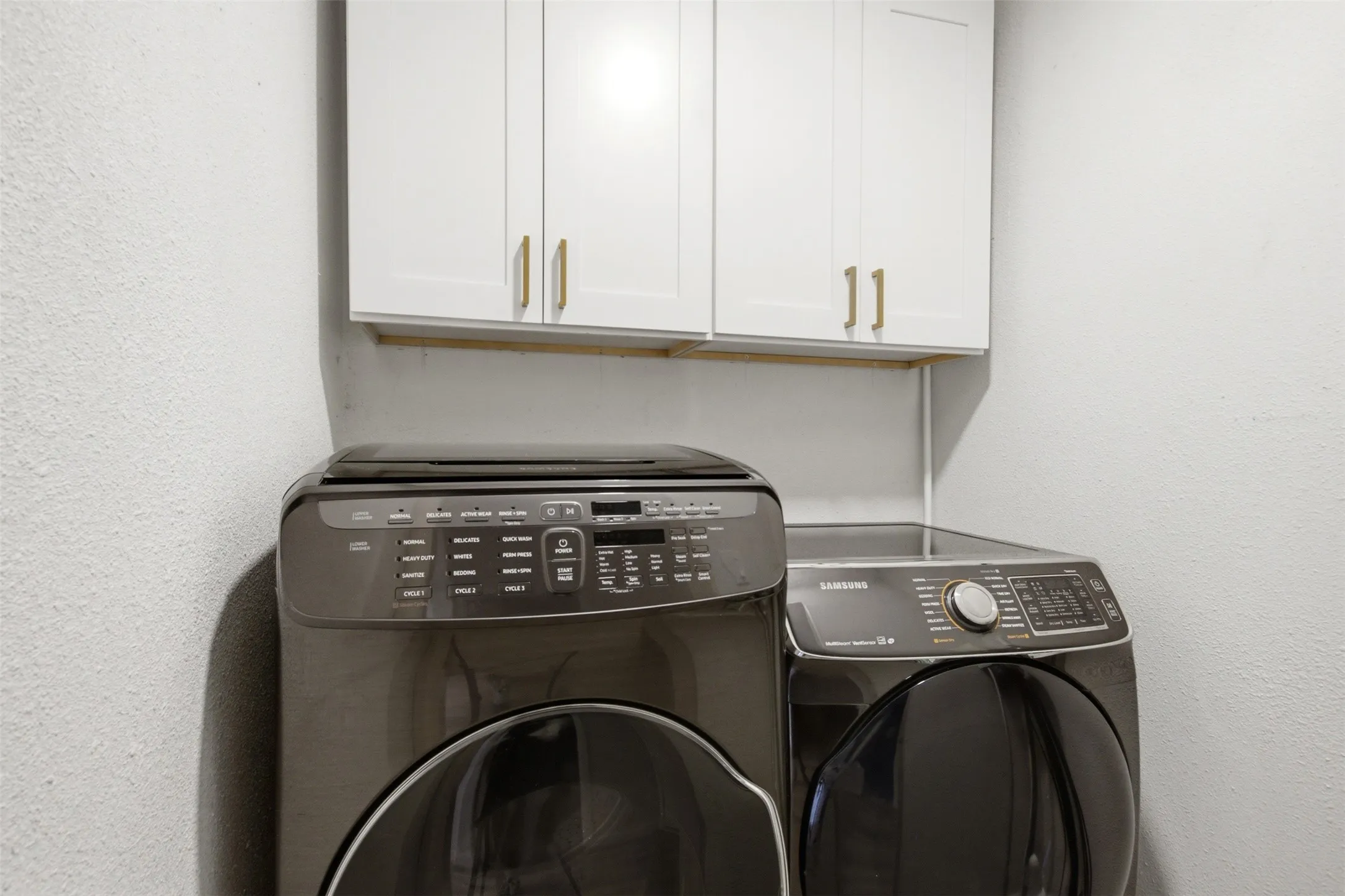 Laundry area featuring white cabinetry with gold-tone hardware, textured walls, and an overhead storage shelf