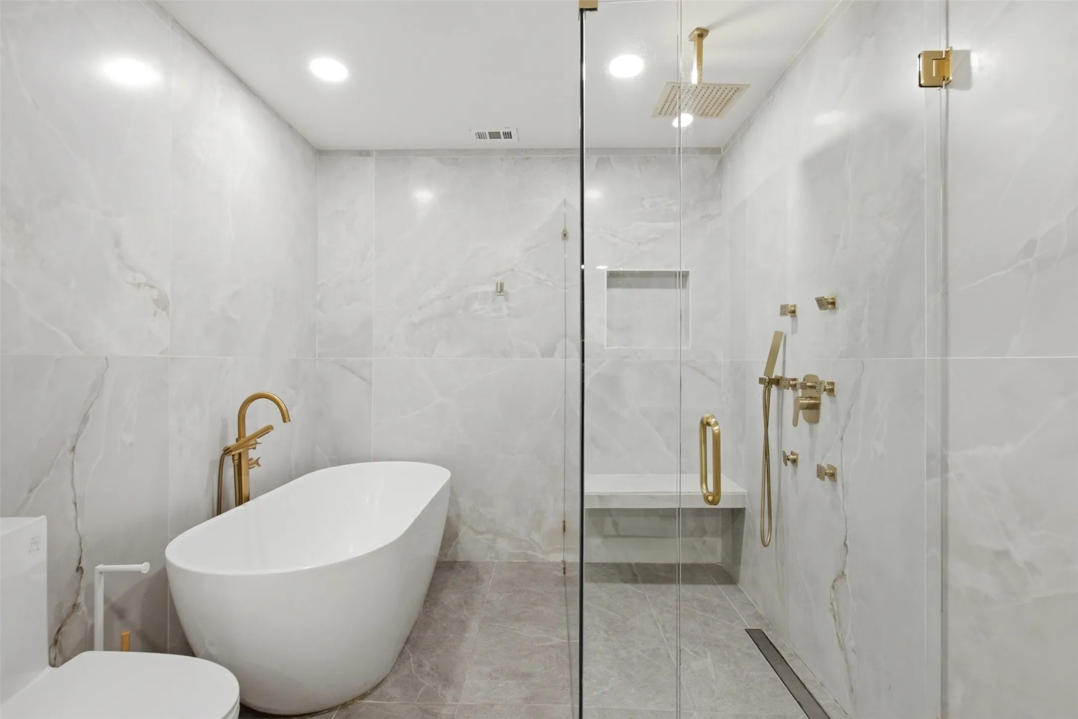 Modern bathroom featuring a freestanding soaking tub with brass fixtures, a glass-enclosed shower with a built-in bench, a rain showerhead, and a handheld shower fixture, all accented by light-toned wall and floor tiling