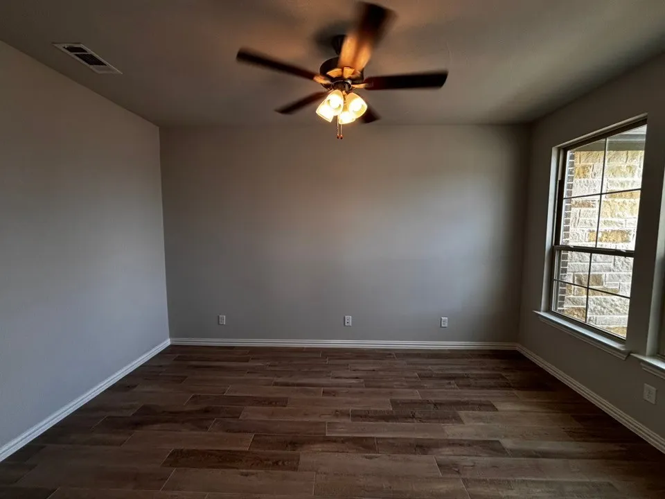 Spacious room featuring wood-finish flooring, neutral wall paint, a ceiling fan with integrated lighting, and a large window providing natural illumination
