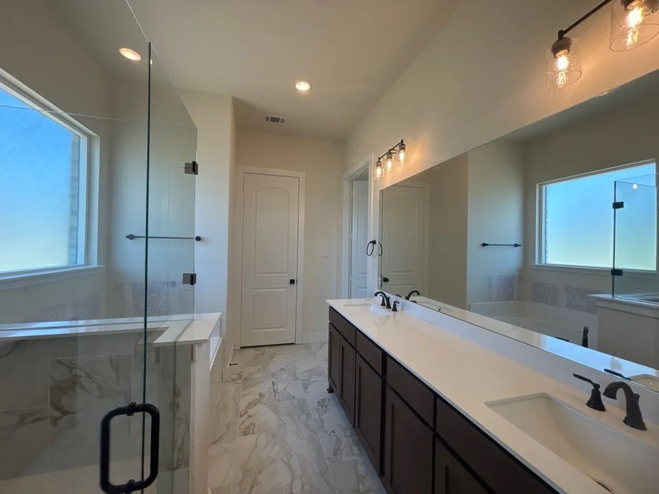 Dual vanity with dark wood-finish cabinetry and white countertops, large wall-mounted mirror, frameless glass shower enclosure, and tiled flooring