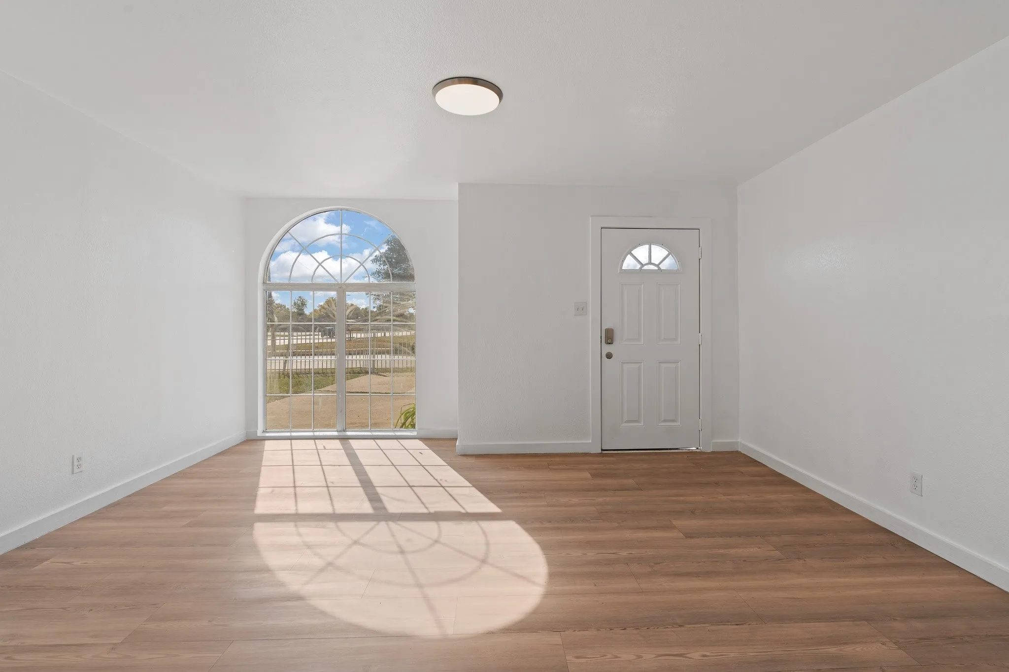Arch-top window, wood-finish flooring, white walls, flush-mount ceiling light, paneled entry door with sunburst lite