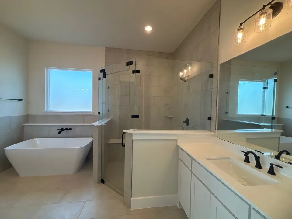 Freestanding soaking tub with a wall-mounted faucet, glass-enclosed shower, dual vanity with an integrated sink, and tiled flooring