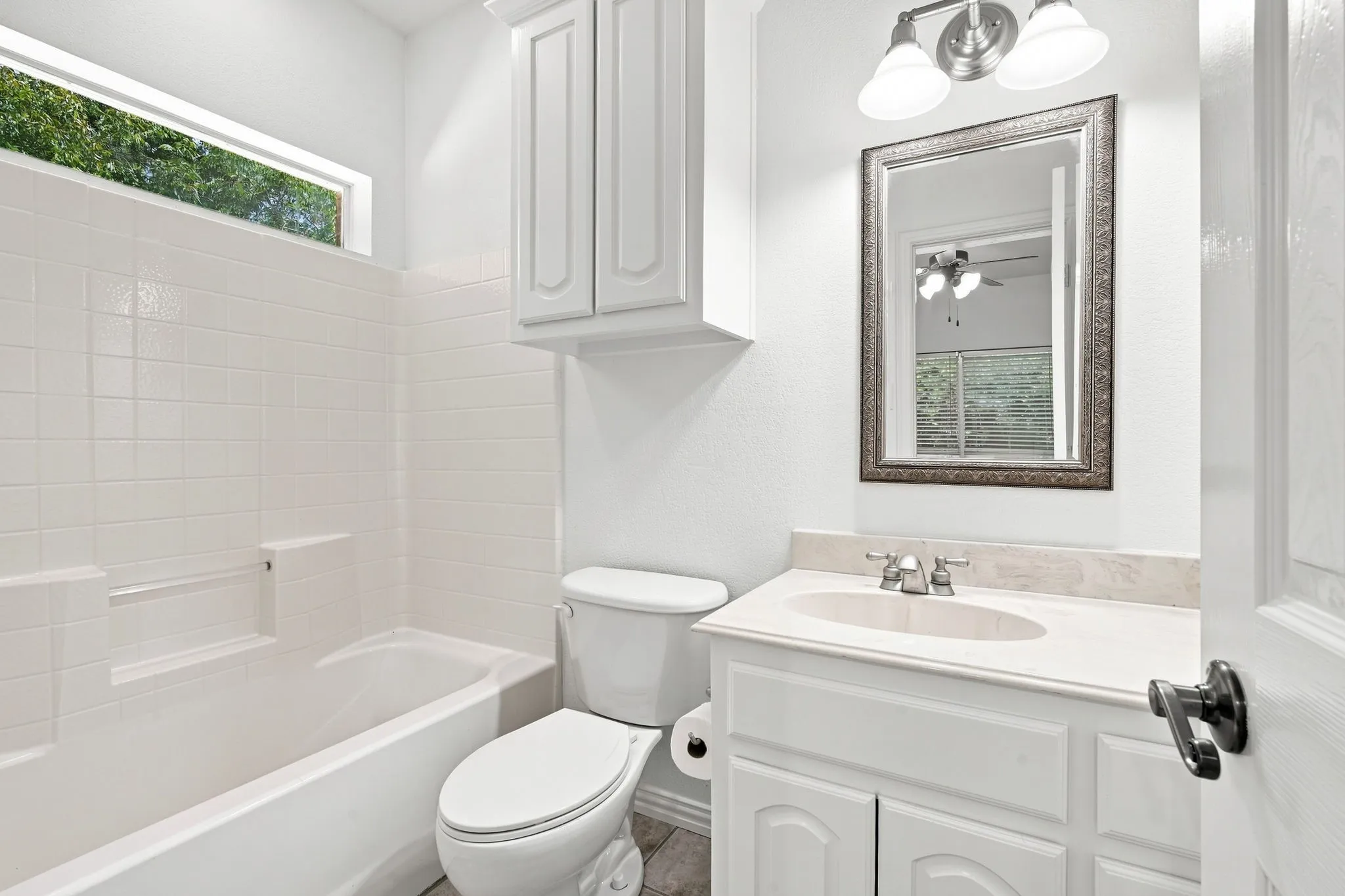 Well-designed bathroom with white ceramic tile surround, a transom window for natural light, and a stone-finish vanity countertop.