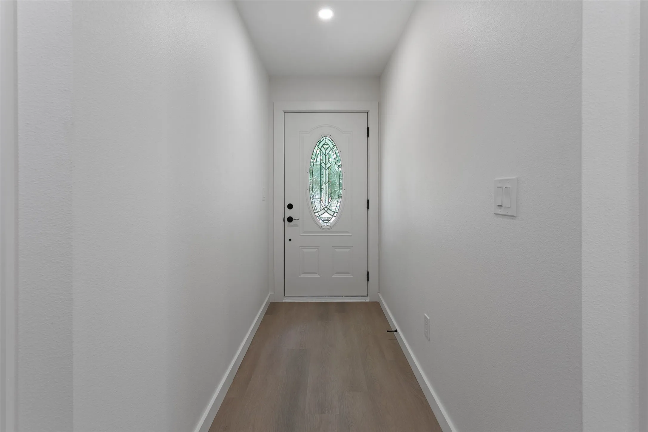 Recessed lighting illuminating the entry with wood-finish flooring, white walls, and a paneled front door with an oval-shaped decorative glass insert