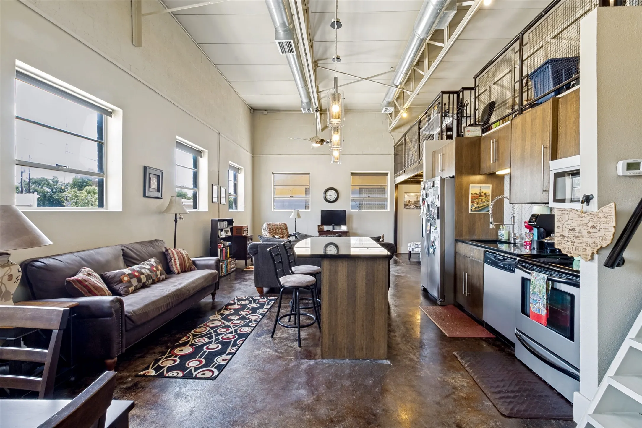 High ceilings with exposed ductwork, concrete flooring, kitchen island with seating, stainless steel appliances, and a mezzanine level with metal railings