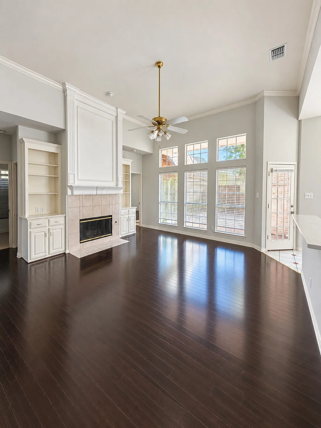 Spacious living area featuring dark wood-finish flooring, a tiled fireplace with built-in cabinetry, large windows, and a ceiling fan with integrated lighting