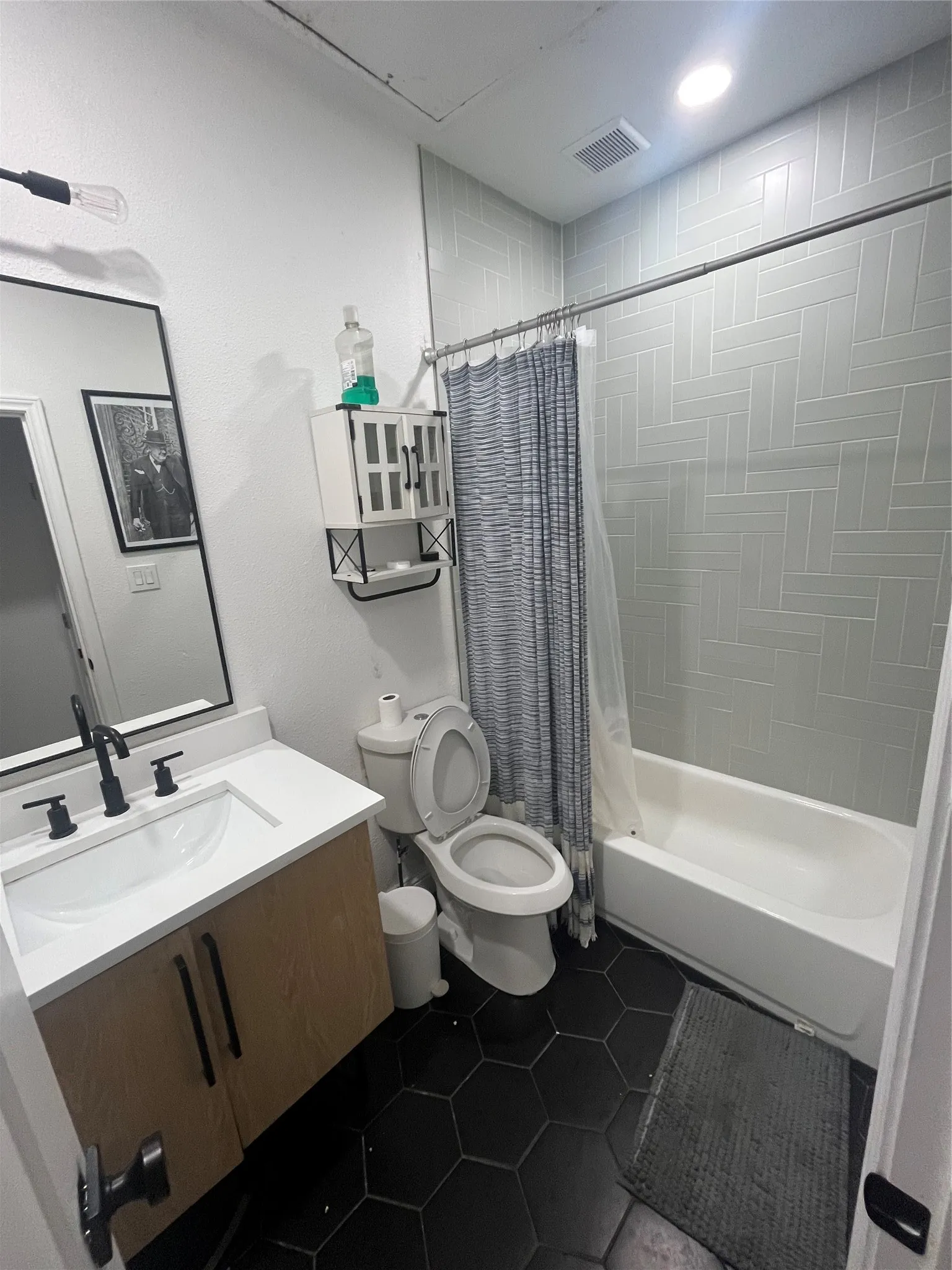 Bathroom featuring a wood-finish vanity with white countertop, black hexagonal floor tiles, and a tub with light-colored subway tile surround