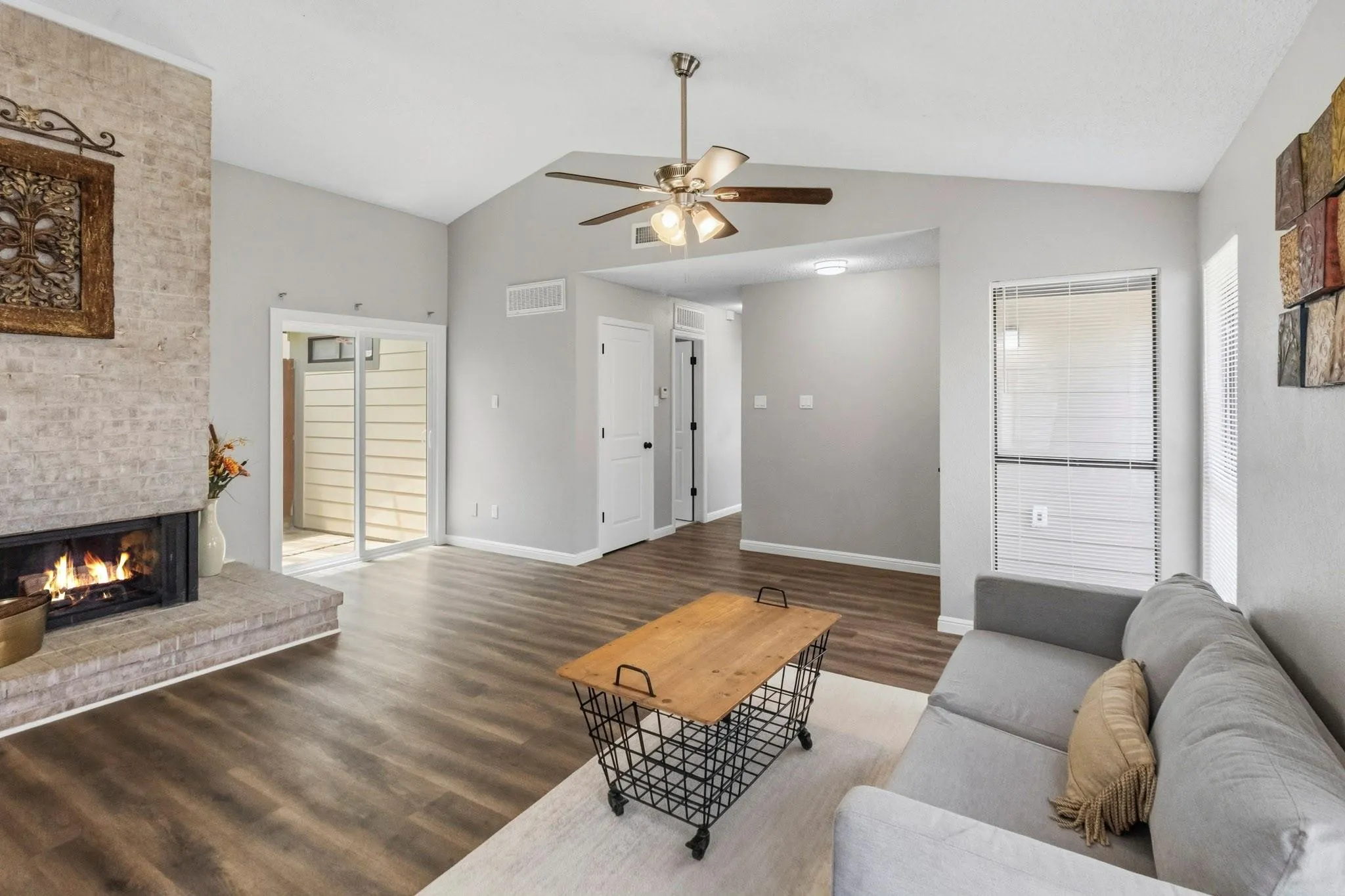 Vaulted ceiling with ceiling fan, brick fireplace, wood-finish flooring, sliding glass door, and neutral wall tones