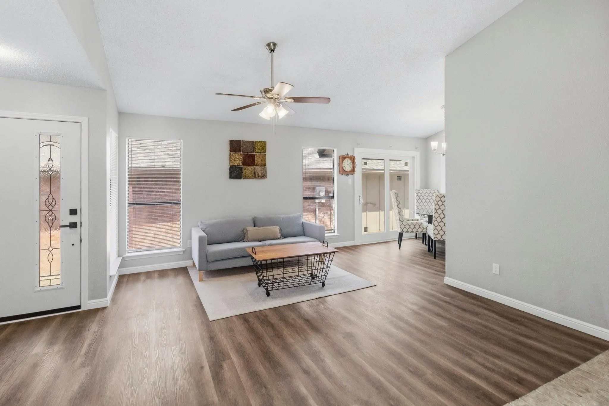 Wood-finish flooring throughout, multiple windows with blinds, ceiling fan, and a white front door with decorative glass inserts