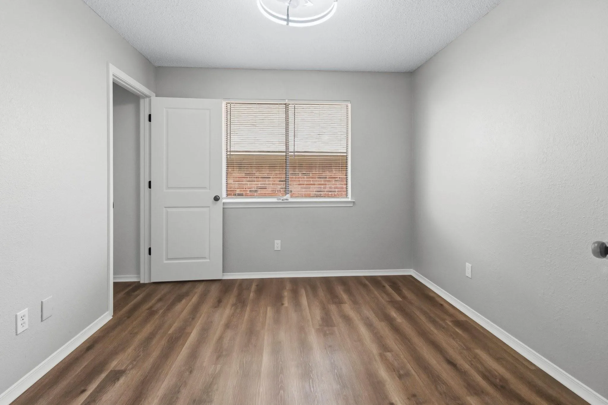 Wood-finish flooring, light gray walls, white trim, single window with blinds, and flush mount lighting