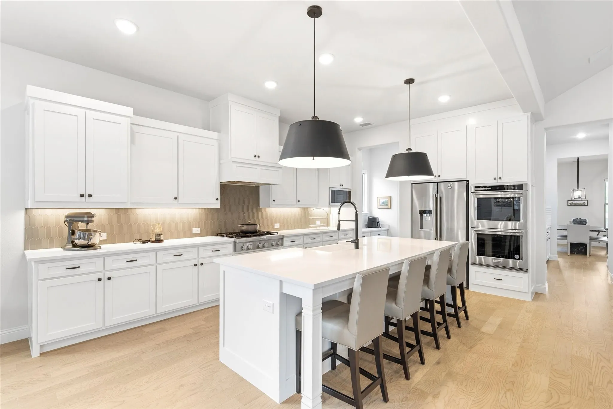 Open Concept Kitchen with white shaker cabinetry, large island, quartzite countertops, tile backsplash, farmhouse sink, stainless steel appliances with double ovens and updated over island lighting.