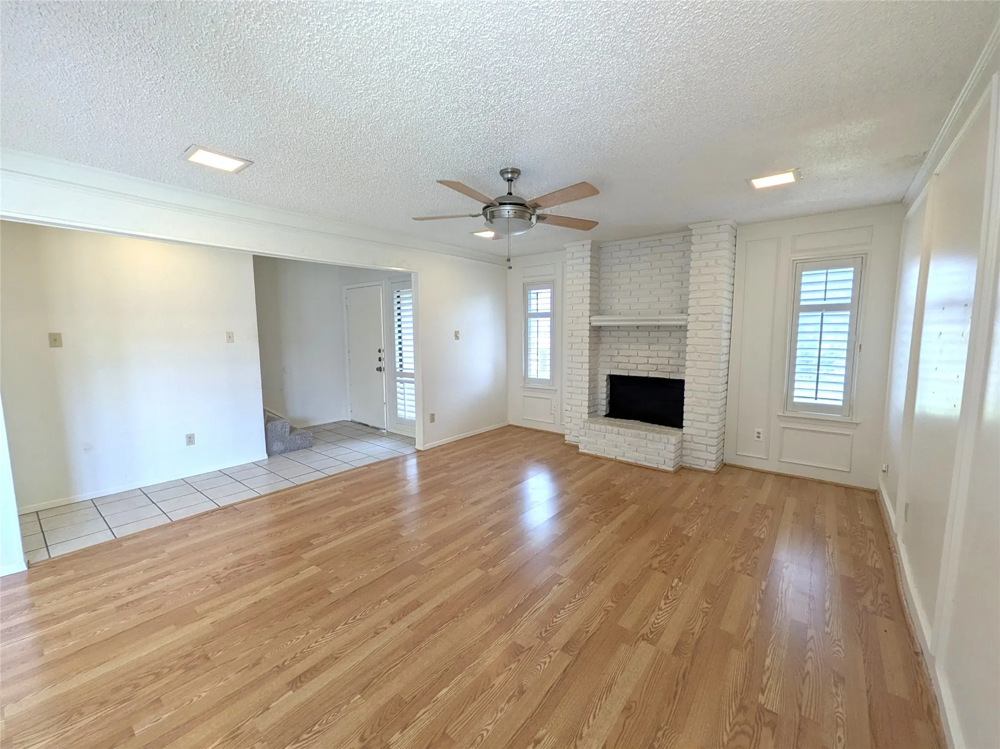 Bright living area featuring wood-finish flooring, a white-painted brick fireplace with a hearth and mantel, and multiple windows with plantation shutters