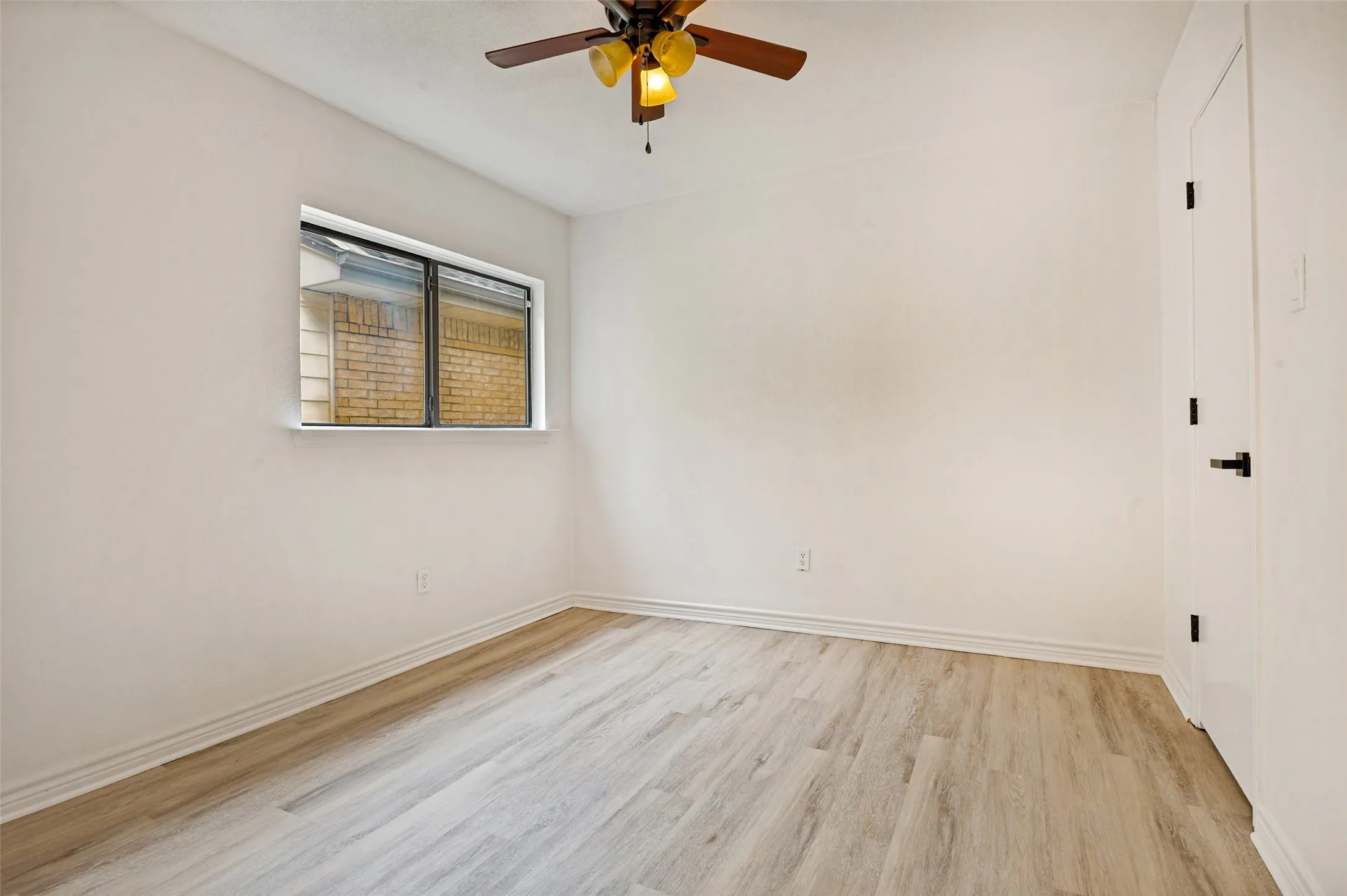 Wood-finish flooring, white walls, rectangular window, ceiling fan with light fixture, and a white door with black hardware