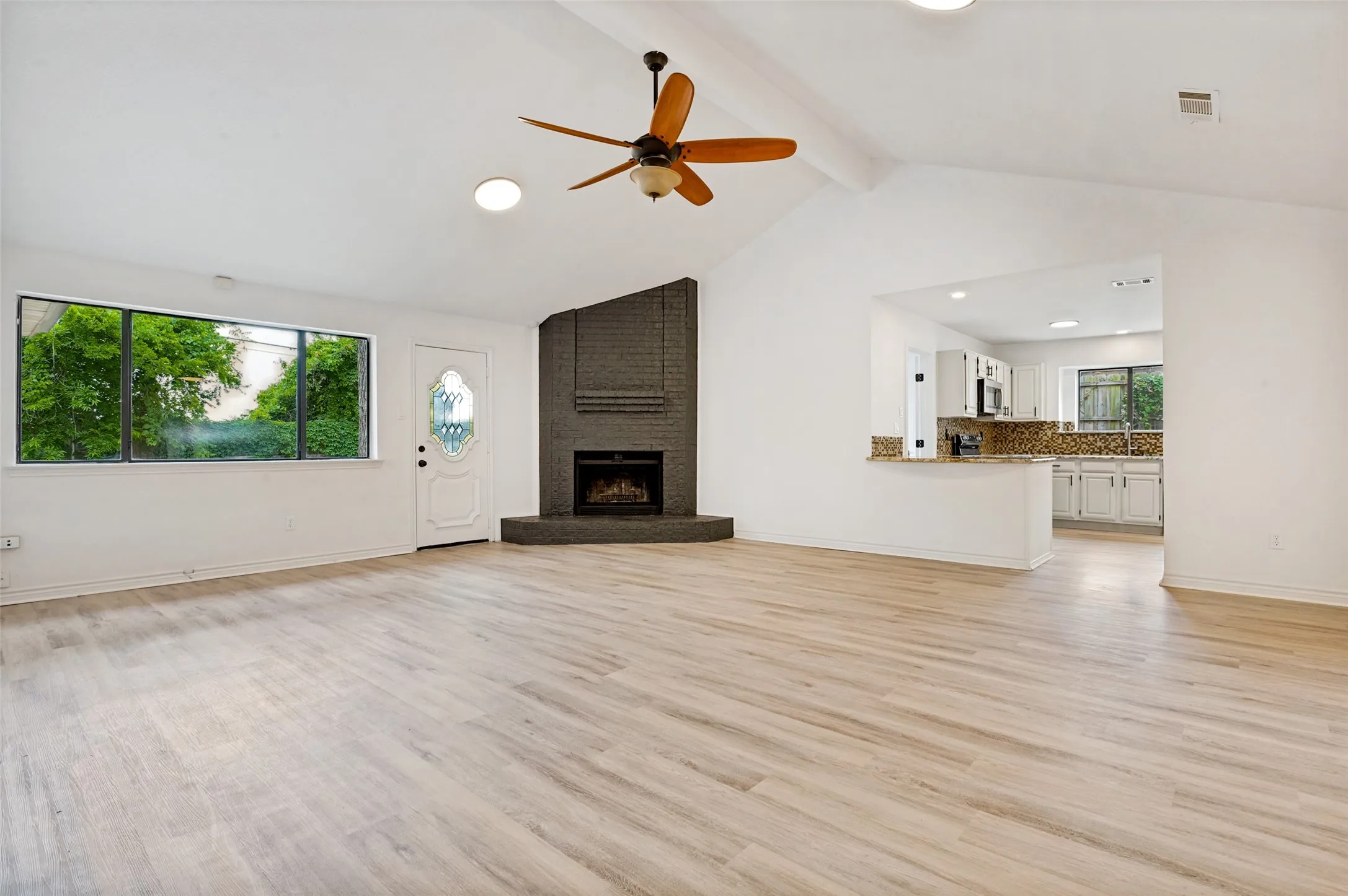 Expansive living area with high vaulted ceilings, light wood-finish flooring, a contemporary ceiling fan, and an accent brick fireplace