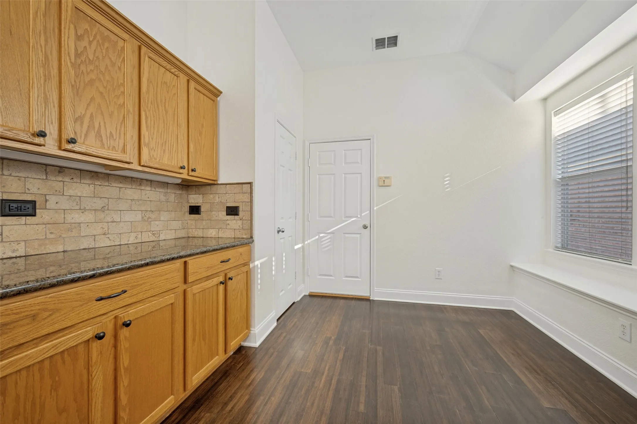 Wood-finish flooring, vaulted ceiling, built-in window bench, light wood cabinetry, and a subway tile backsplash