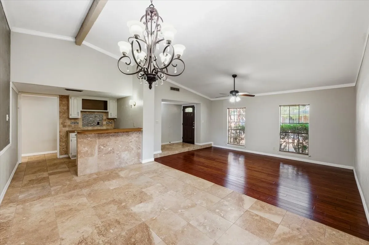 Dining Area features a vaulted ceiling with exposed wood beam and a chandelier light fixture, opening to a built-in butlers pantry with tile backsplash and views through to the living room and front entry. Tile flooring transitions to wood in the connected living space.