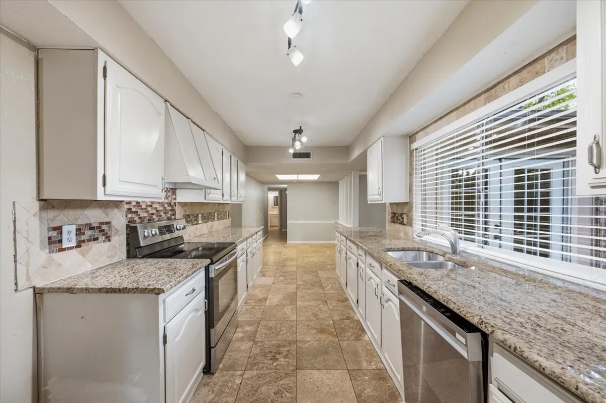Kitchen features track lighting along the galley layout, with granite countertops and tile backsplash. A built-in dishwasher sits beside the sink beneath a window, and the space opens toward the hallway and breakfast area. Tile flooring extends throughout.