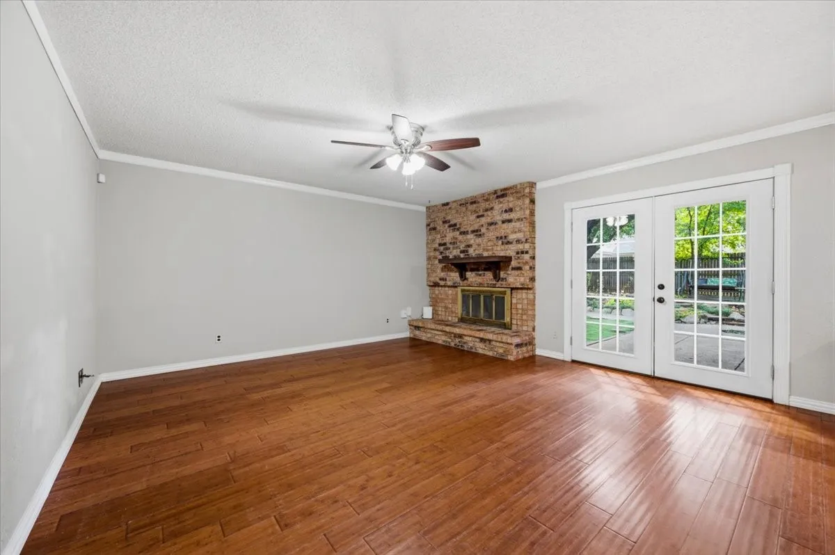 Family Room features crown molding and ceiling fan with light fixture, centered on a brick wood-burning fireplace with wood mantle. French doors open to the backyard. Wood flooring extends throughout.