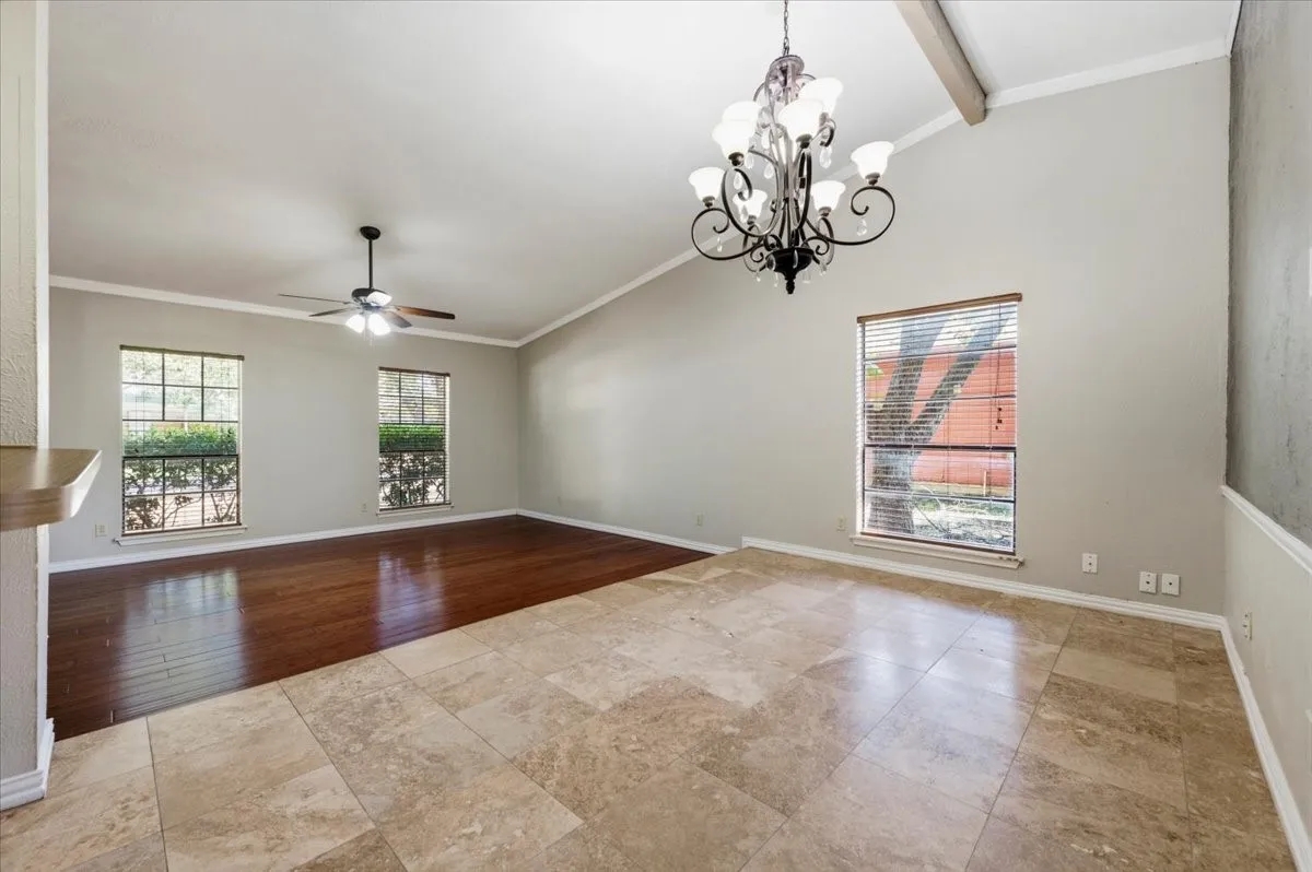 Dining Area features the same vaulted ceiling and exposed wood beam from the opposite angle, with two windows bringing in natural light alongside the chandelier overhead. Tile flooring transitions to wood in the connected living space.