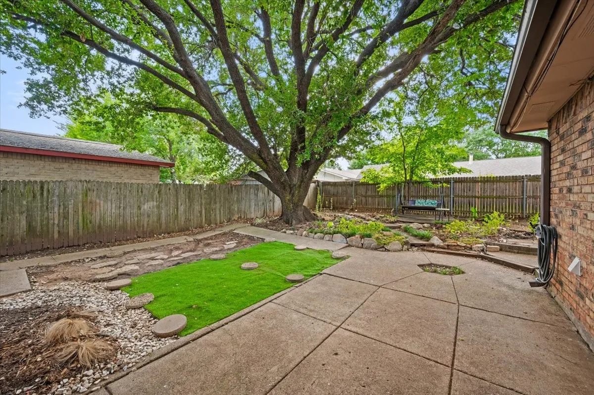 Backyard features a concrete walkway, a mature shade tree, wood privacy fencing, and a landscaped area with decorative stone accents.