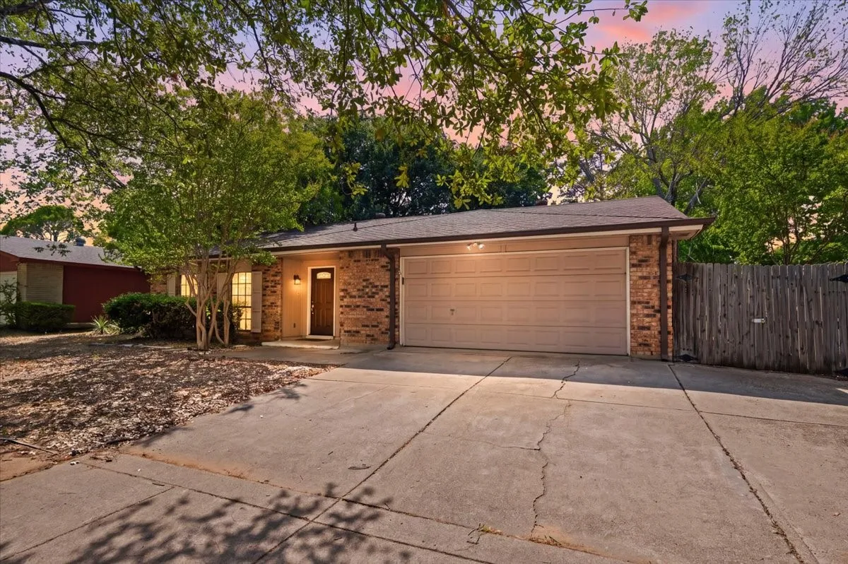 Front Exterior features a brick and wood exterior with an attached two-car garage, concrete driveway, and covered front entry. Mature trees frame the home, with wood privacy fencing along the side yard.