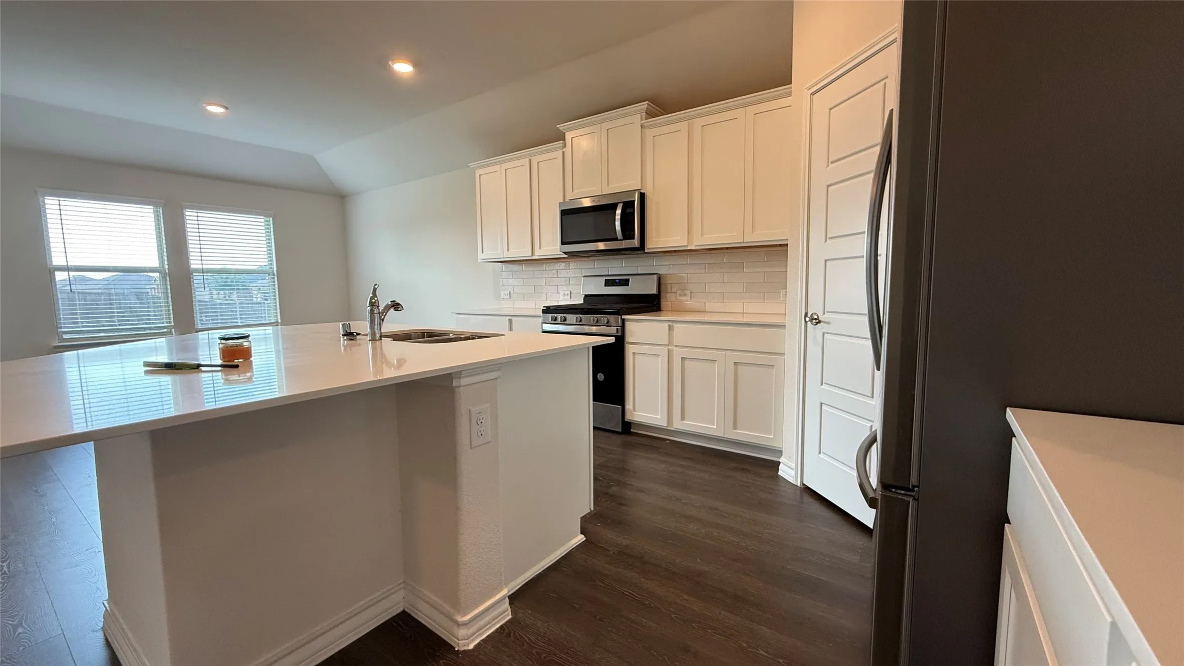 Kitchen featuring a central island with an integrated sink, stainless steel appliances, white cabinetry, tile backsplash, and dark wood-finish flooring