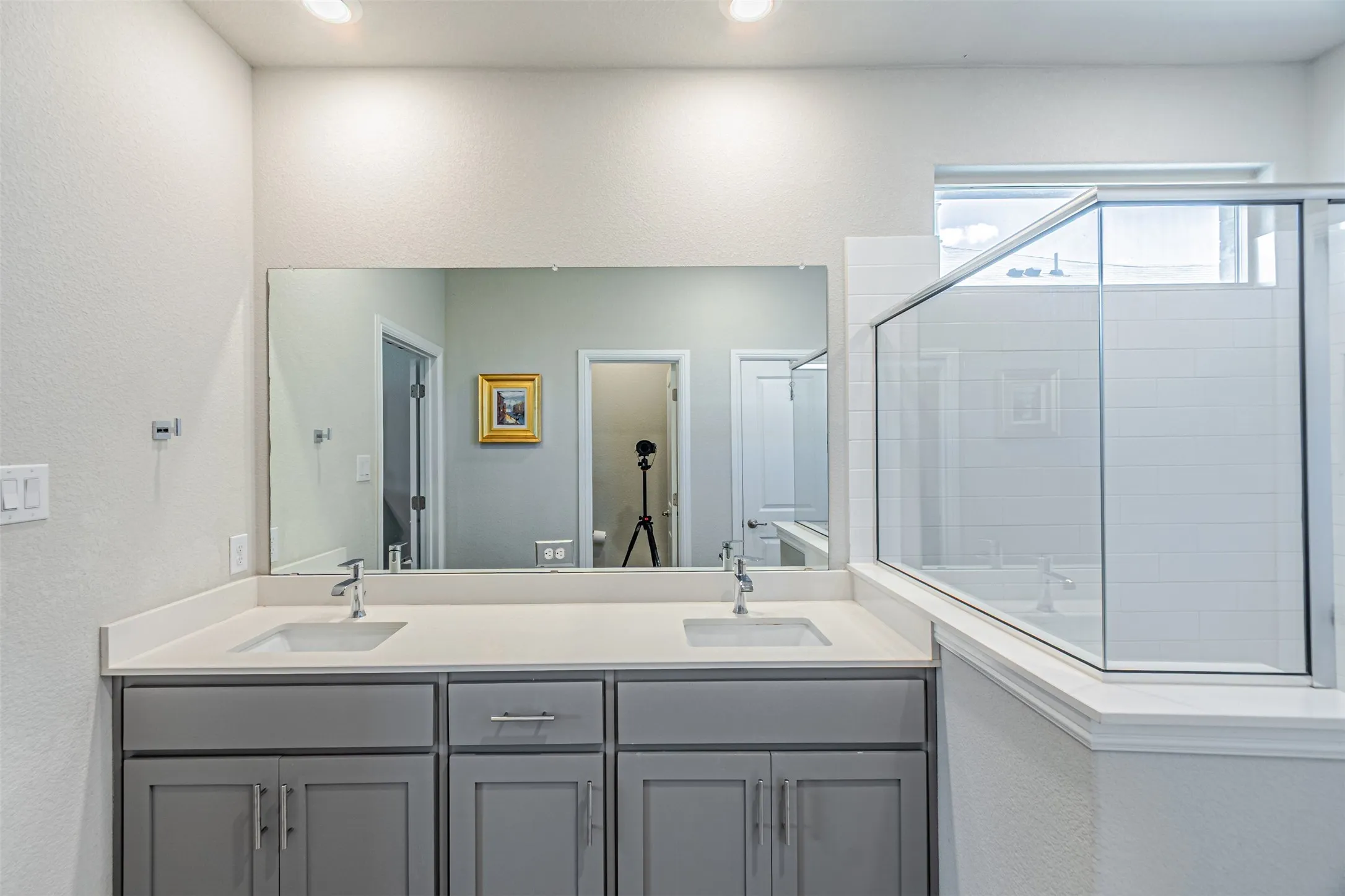 Dual sink vanity with light gray cabinetry and chrome hardware, a large wall-mounted mirror, and a glass-enclosed shower featuring white subway tile