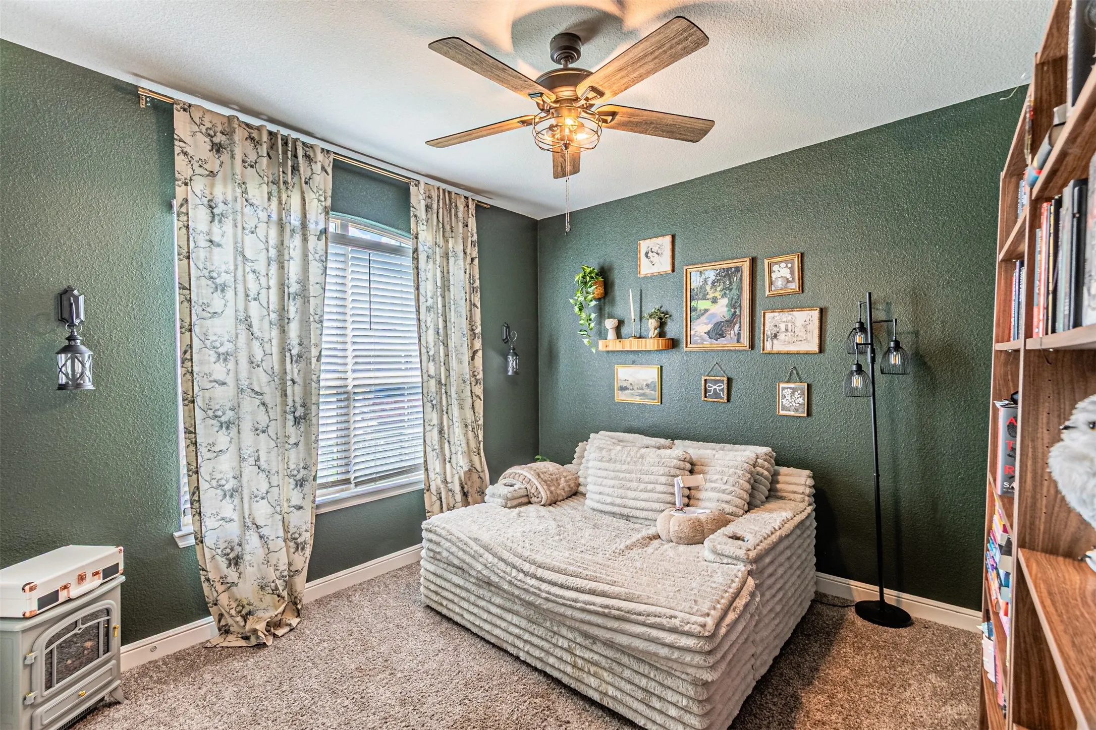Textured green walls, ceiling fan with integrated lighting, window with blinds, carpeted flooring, and a built-in wood-finish bookshelf