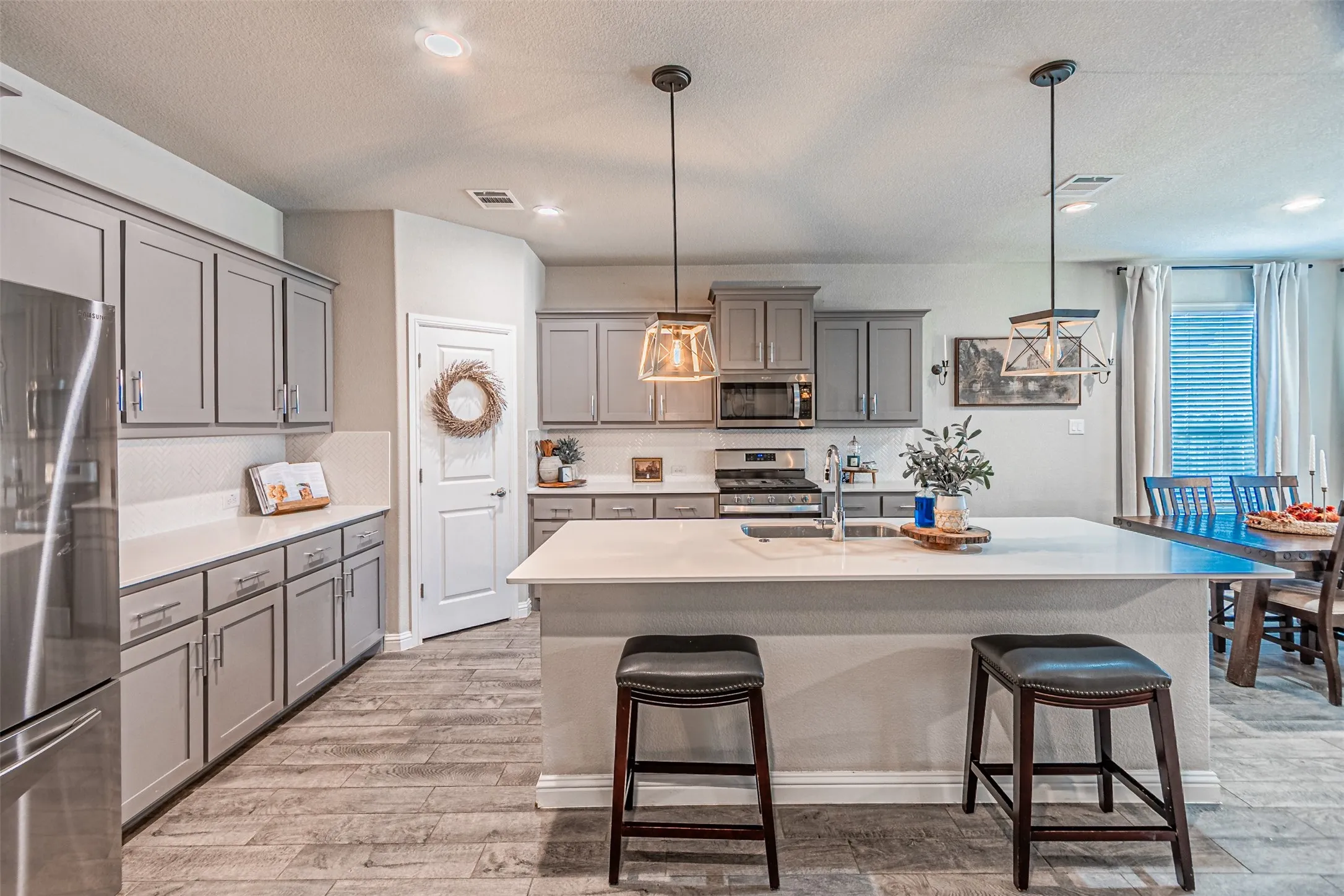 Kitchen featuring wood-finish flooring, light gray cabinetry, stainless steel appliances, a white tile backsplash, and a kitchen island with a built-in sink