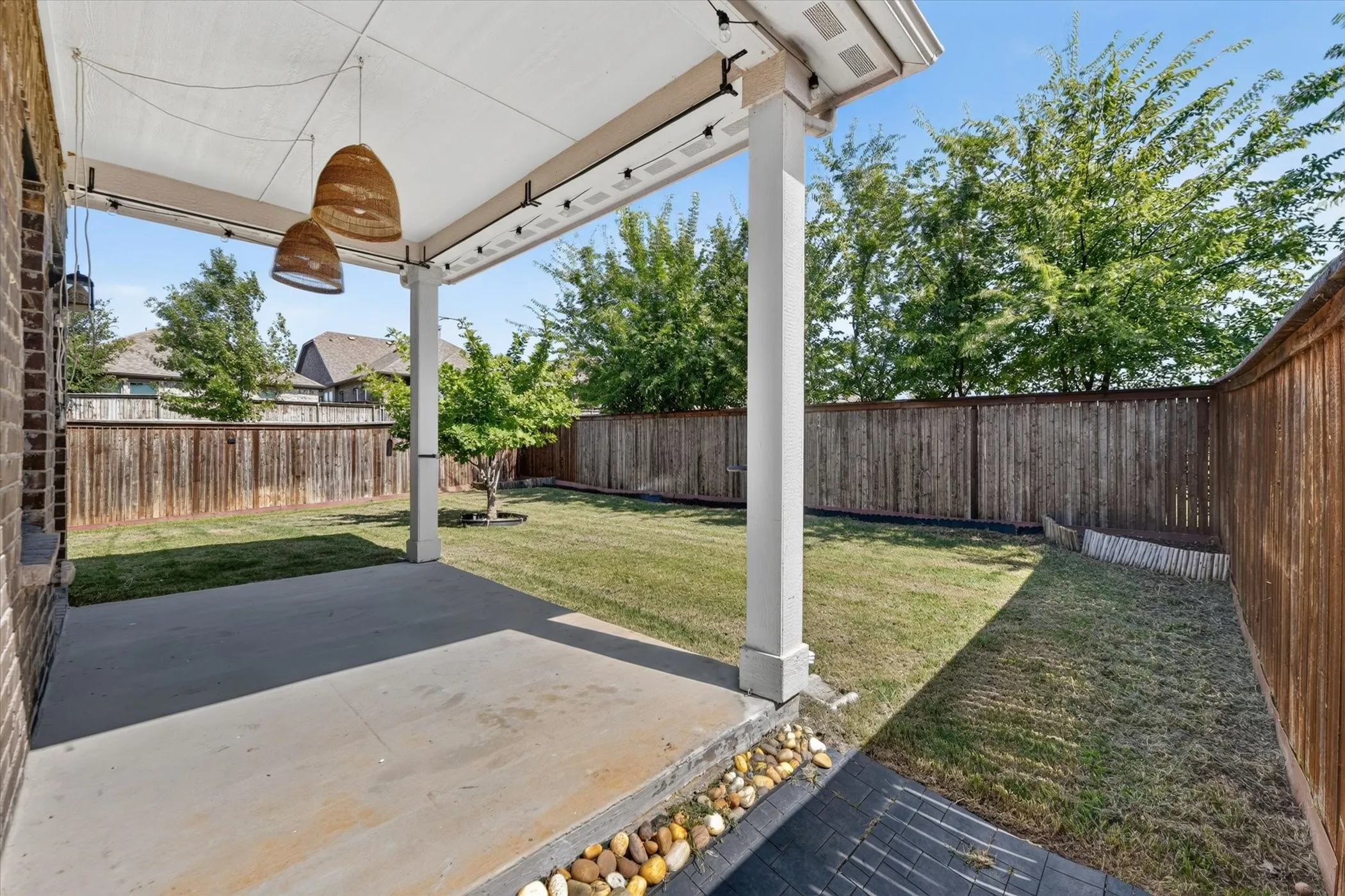 Covered patio with concrete flooring, two hanging light fixtures, and a fenced backyard with a small tree