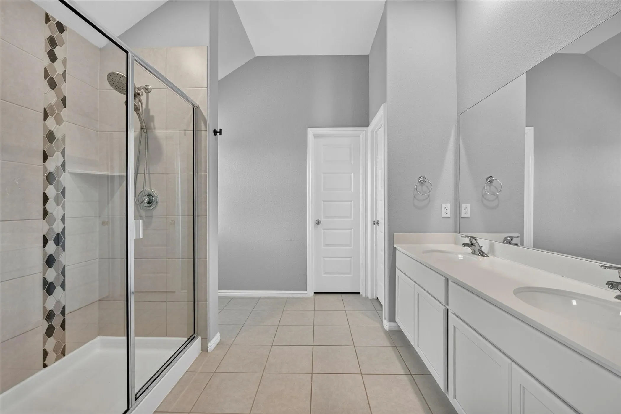 Dual vanity with integrated sinks and white cabinetry, large wall-mounted mirror, tiled flooring, and a shower with glass enclosure