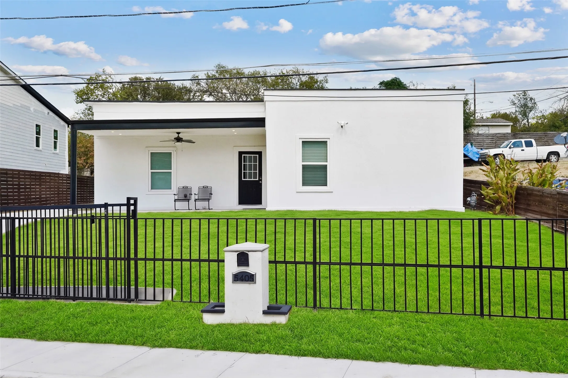 White exterior with a flat roofline, covered front patio featuring a ceiling fan, black entrance door, and a black metal fence enclosing the front yard