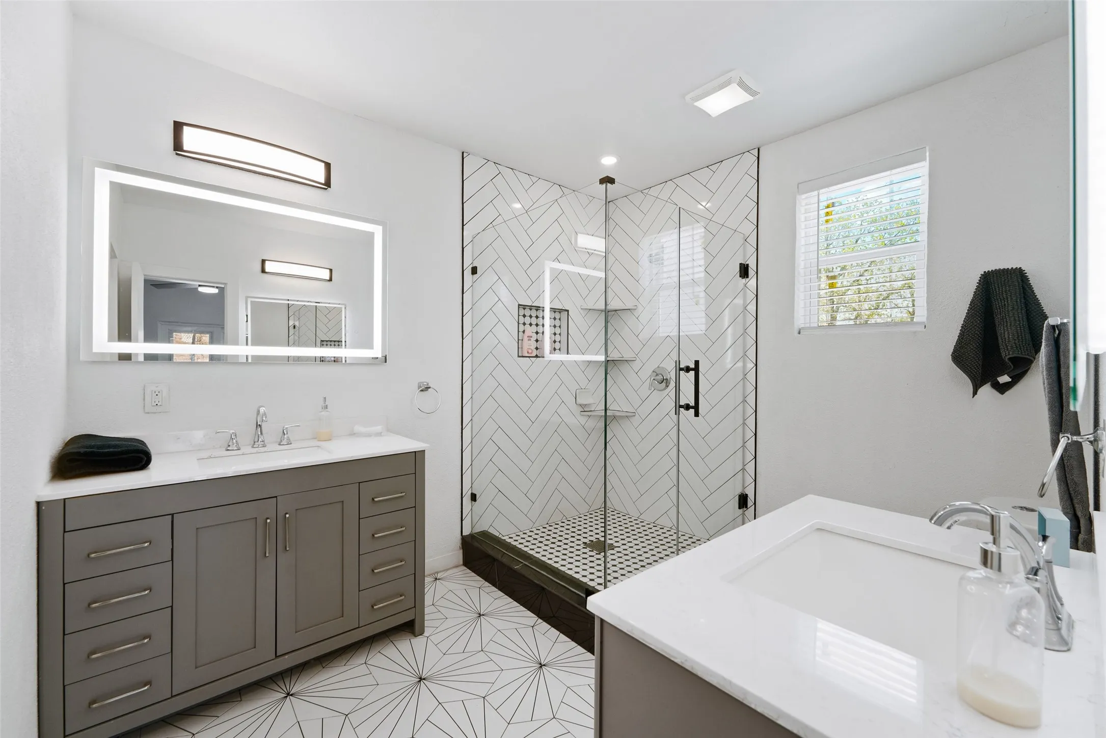 Dual vanities with white countertops, a frameless glass shower enclosure, white subway tile with a herringbone pattern, and patterned floor tiling