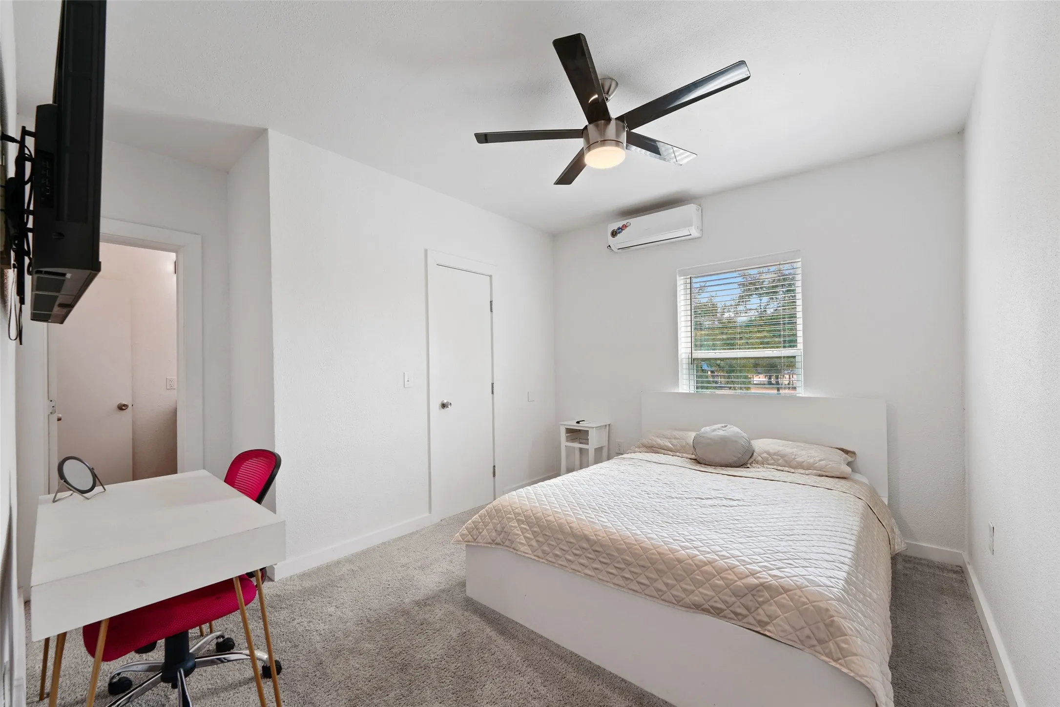 White interior walls, ceiling fan with integrated lighting, window with horizontal blinds, wall-mounted air conditioning unit, and plush grey carpeting